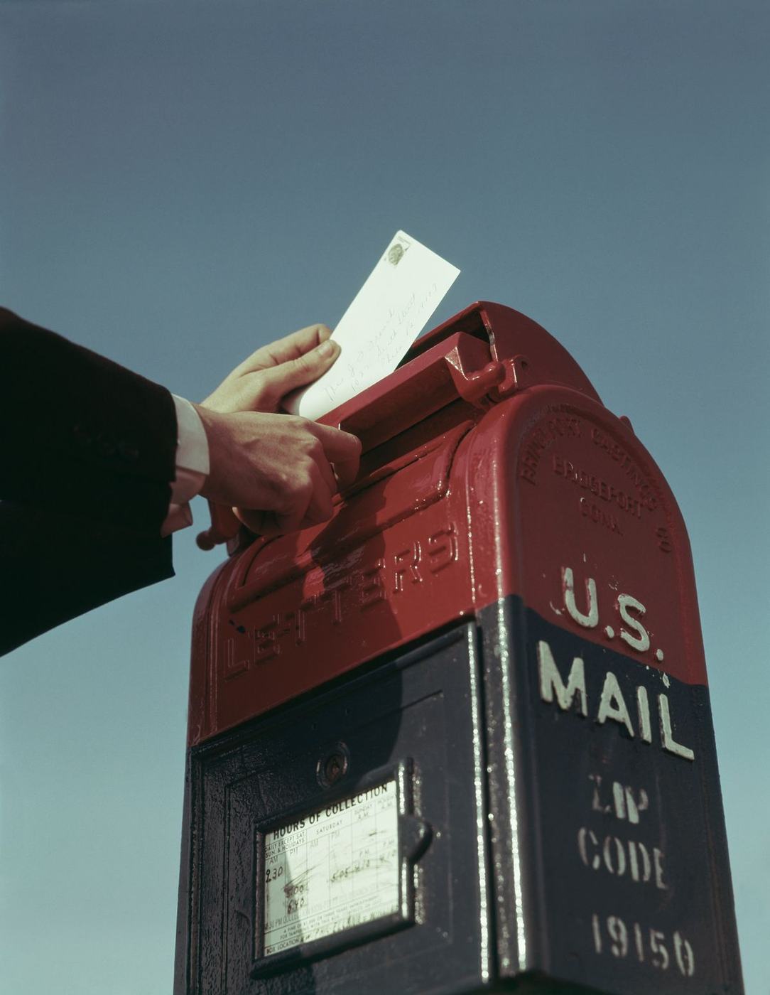 Hand posting a letter into a red and navy US mailbox